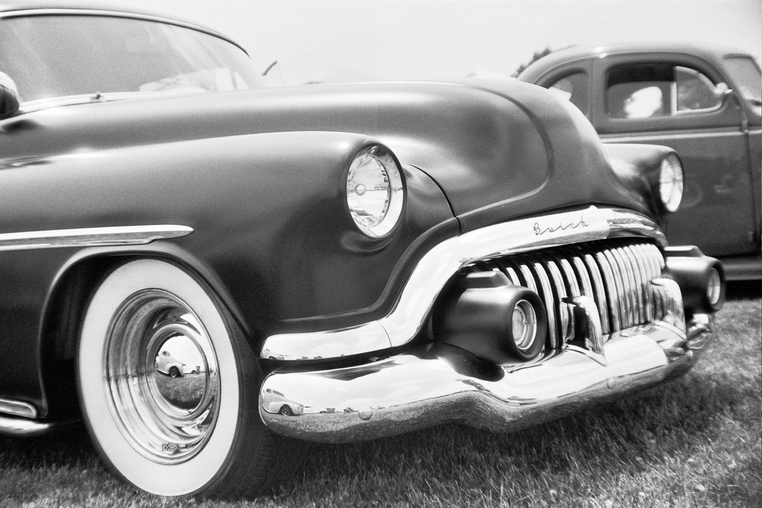 Black and white close-up photo of a classic Buick car. The car features large round headlights, a prominent chrome grille with vertical bars, chrome bumper accents, and whitewall tires. Another vintage car is partially visible in the background—perfect inspiration for Millworks Web Design & Graphics.