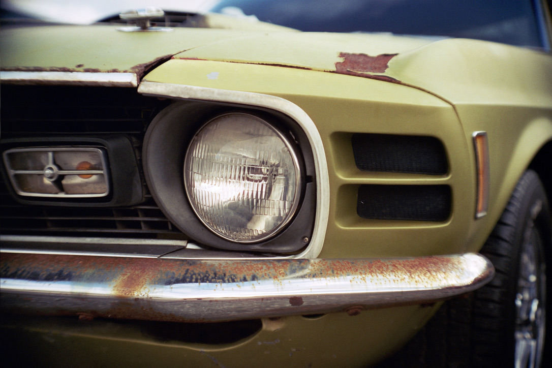 Close-up of the front of an old, weathered car with a greenish-yellow paint job. The car's headlight, grill, and part of the bumper are visible. There are signs of rust and peeling paint, giving the car a vintage and aged appearance—much like a classic aesthetic in web design by Millworks Web Design & Graphics.