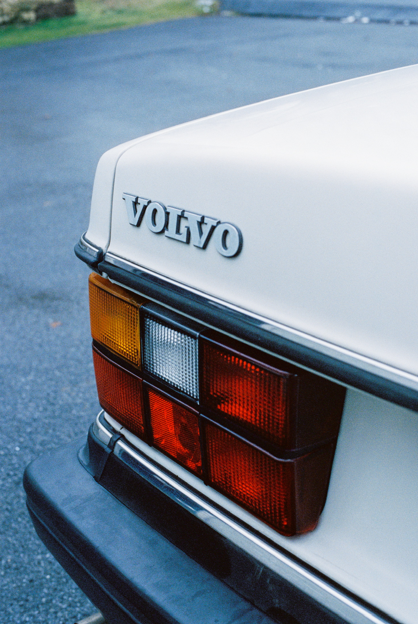 Close-up of the rear corner of a white Volvo car, showing the brand logo and the taillight design including red, amber, and white lights. Set against a background of asphalt and greenery, the image evokes precision much like Millworks Web Design & Graphics' custom websites. The trunk and rear bumper are also partially visible.