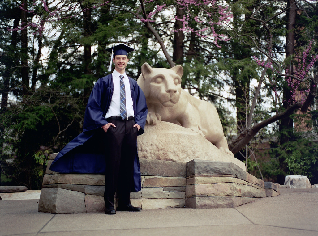 A person wearing a blue graduation gown and cap stands next to a stone statue of a reclining lion. The background features green trees and pink flowering branches. The person is smiling, holding the gown open with one hand, reminiscent of showcasing their talent in graphic design.