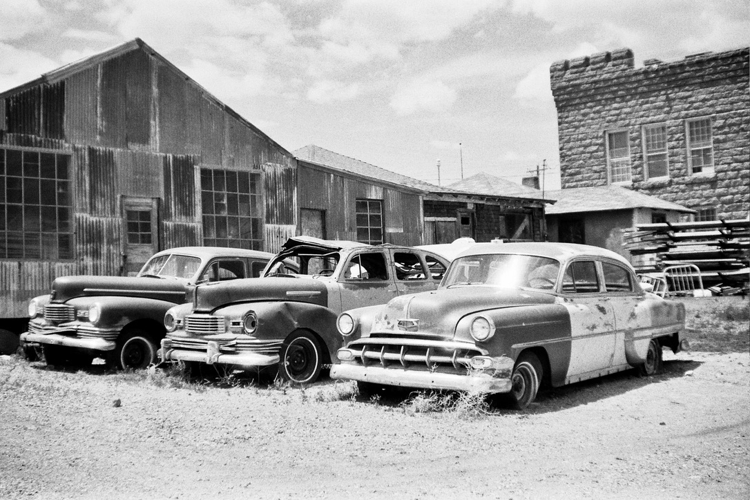 A black-and-white photo shows three classic cars parked in front of an old, weathered building with corrugated metal siding and a stone structure. The cars appear worn and rusted, evoking a sense of nostalgia and the passage of time—much like the story behind Millworks Web Design & Graphics' custom websites.