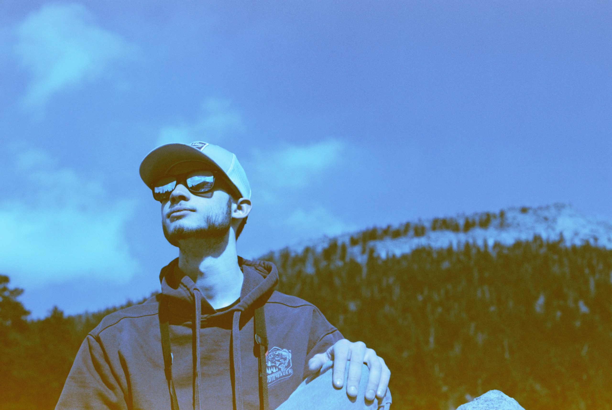 A person wearing a baseball cap and sunglasses stands outdoors against a mountain backdrop. The image has a blue tint giving it a vintage feel, reminiscent of classic graphic design. The individual, dressed casually in a hoodie, seems to be looking off into the distance.