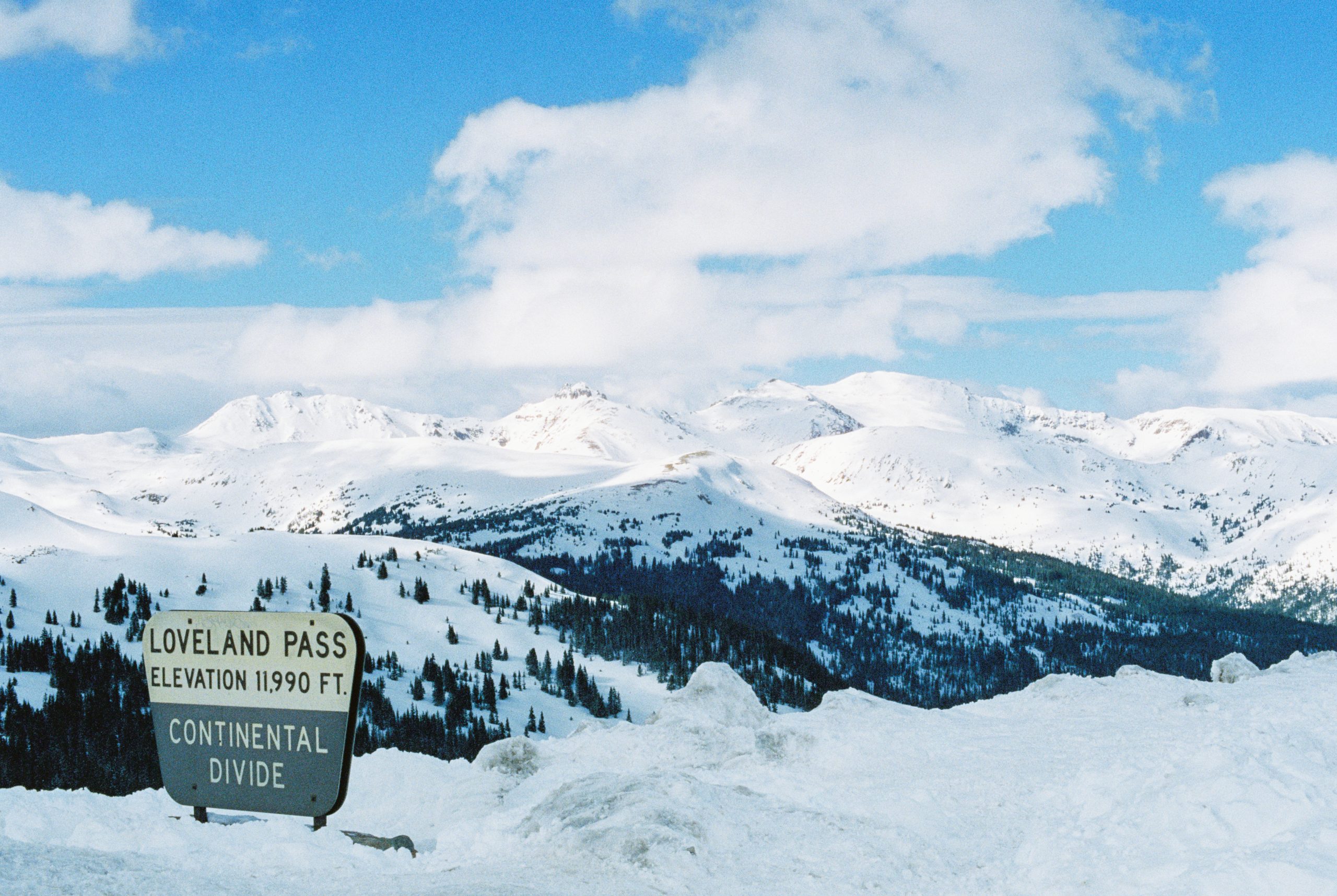 A scenic view of Loveland Pass, featuring snow-covered mountains under a partly cloudy sky. A sign in the foreground reads "Loveland Pass Elevation 11,990 ft. Continental Divide." Snow blankets the terrain, crafting a pristine, wintry landscape perfect for graphic design inspiration or digital marketing campaigns.