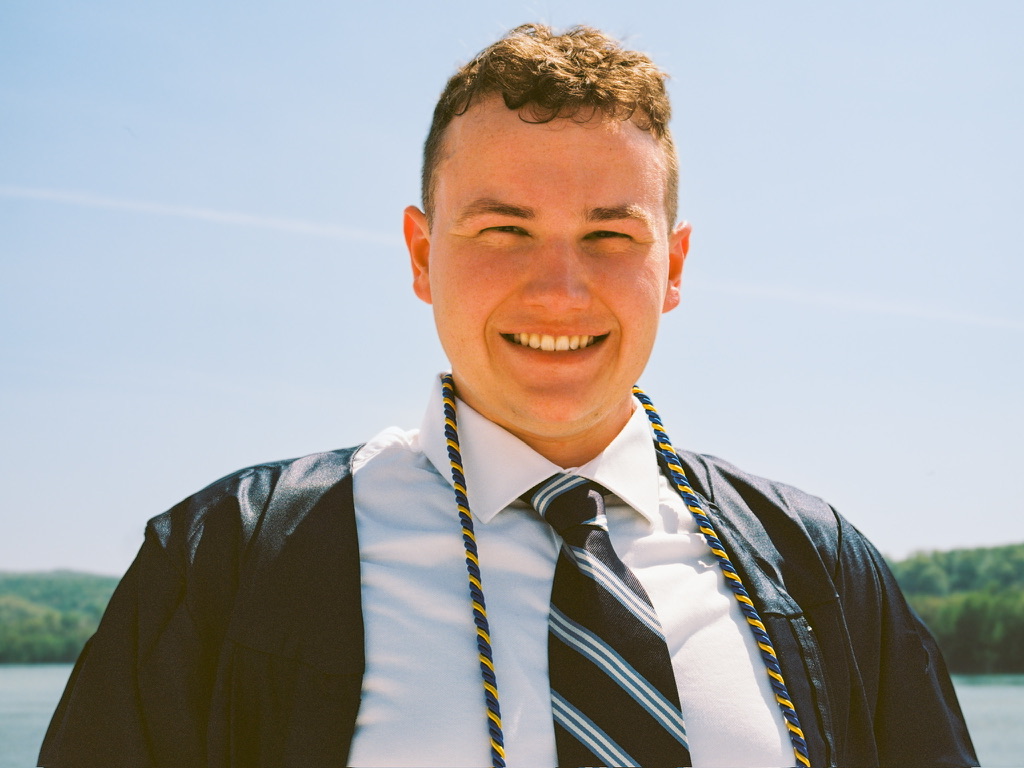 A smiling young man wearing a graduation cap and gown stands outdoors with a scenic view of water and trees behind him. Adorned with cords indicating academic achievement in graphic design, he basks under the clear blue sky of a sunny day.