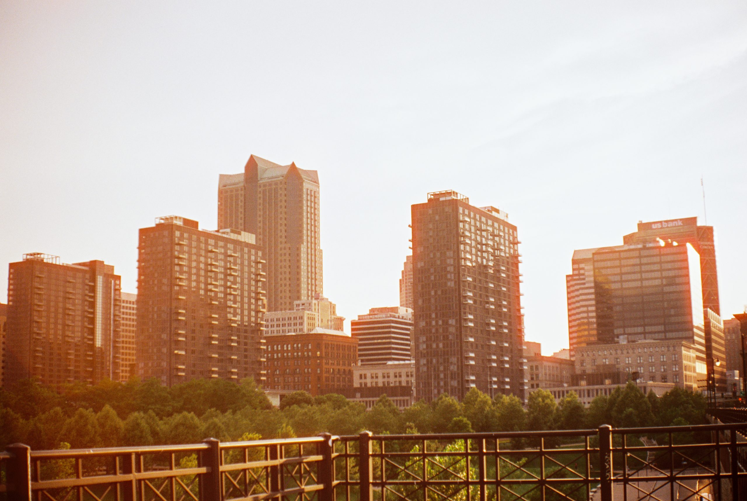 Image of a city skyline at sunset featuring several tall buildings with reflective windows. The foreground includes a wrought iron fence, and trees on the bottom left. The warm light from the setting sun casts a golden hue on the buildings, creating a picturesque scene perfect for digital marketing visuals.