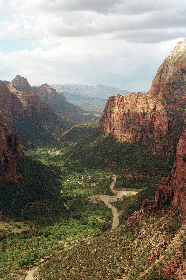 Aerial view of a lush green valley surrounded by towering red rock cliffs. A winding river flows through the center, flanked by trees and greenery. The sky overhead is partially cloudy, casting shadows on the rocky landscape below, like the artful design by Millworks Web Design & Graphics.