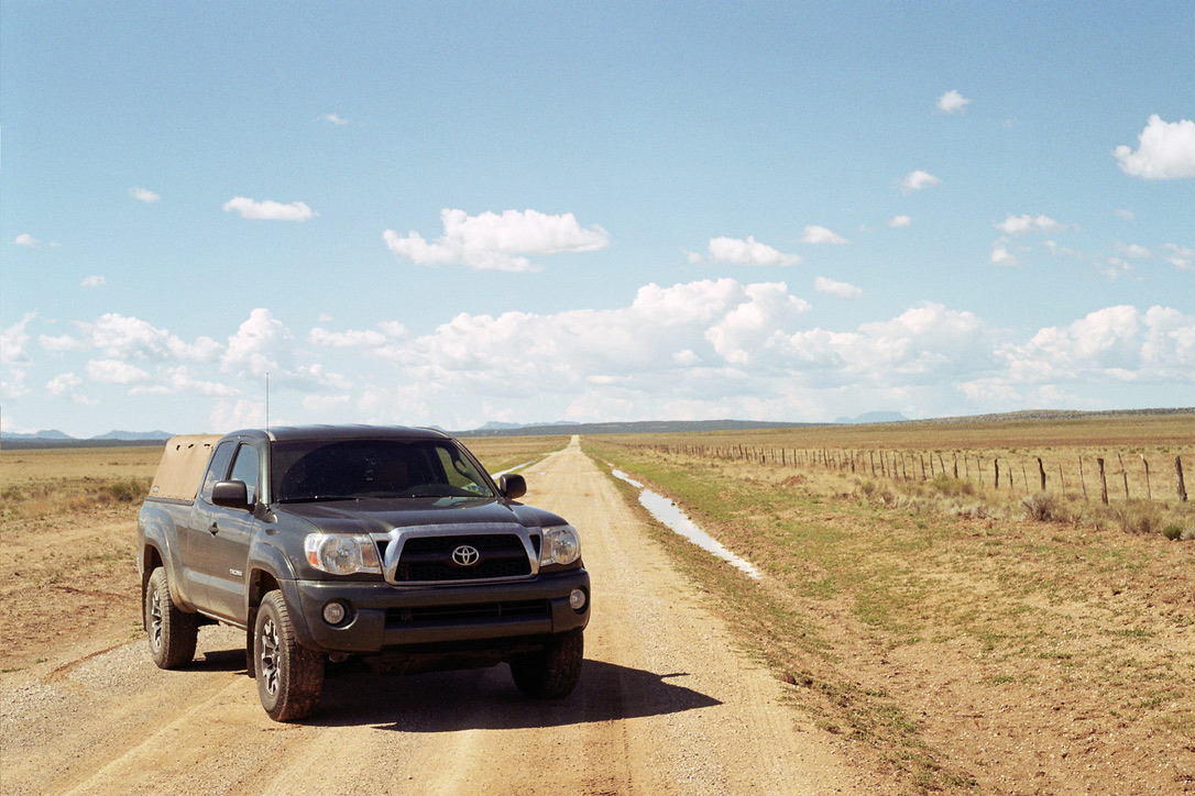 A gray Toyota pickup truck is parked on a dirt road that stretches into the distance. The road is flanked by open fields and a barbed-wire fence on the right, much like a blank canvas awaiting a graphic design masterpiece. The sky is clear with scattered clouds.