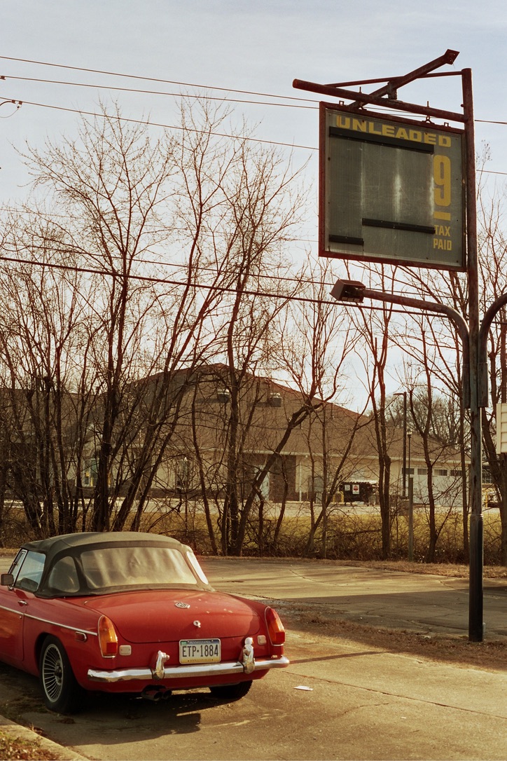 A parked red vintage convertible car with a black soft top is in the foreground. The license plate reads "EP-1884." In the background, an old, weathered gas price sign partially obscured by leafless trees displays "UNLEADED 9." This scene could be perfect for a Millworks Web Design & Graphics project.