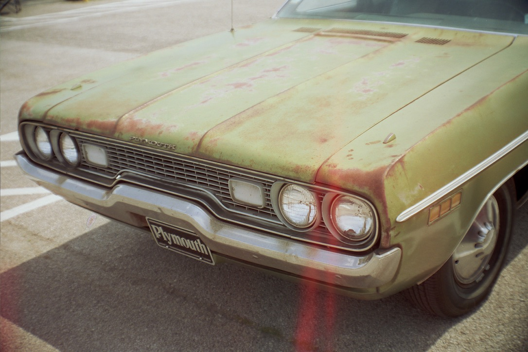 A vintage green Plymouth car with a rusty and weathered exterior is parked on a concrete surface. The front grille and headlights are partially visible, along with the Plymouth logo on the bumper. This classic vehicle's timeless design could effortlessly inspire custom websites for Millworks Web Design & Graphics.