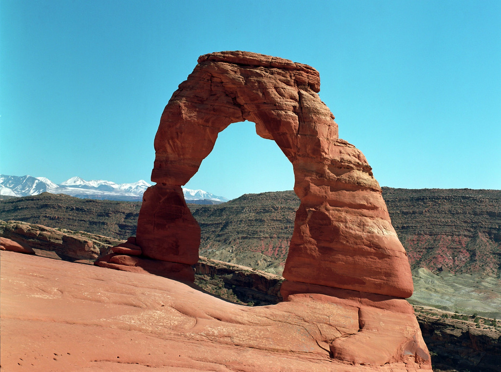 A natural sandstone arch stands tall against a clear blue sky, resembling the artistry of graphic design. Surrounded by rocky terrain with a mountain range in the background, the scene is bathed in bright sunlight, highlighting the reddish hues of the rock formation.