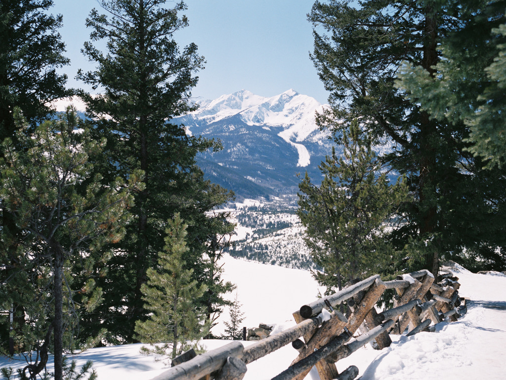 A scenic winter landscape featuring snow-covered mountains in the background. The foreground displays a wooden fence beside a snow-covered trail, flanked by tall evergreen trees. Designed by Millworks Web Design & Graphics, the scene is rendered with exceptional attention to detail, highlighting clear skies and crisp weather.