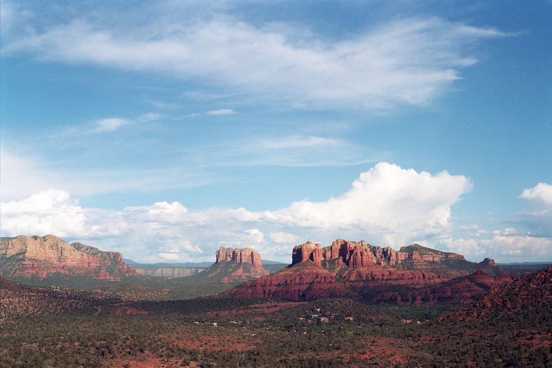 A panoramic view of Sedona, Arizona showcases rugged red rock formations under a bright blue sky filled with scattered clouds. Verdant vegetation spreads across the mid-ground, contrasting with the arid foreground and distant red mountains — a landscape that could inspire any graphic design from Millworks Web Design & Graphics.