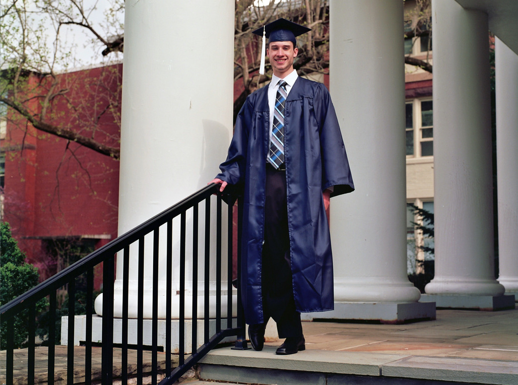 A man wearing a blue graduation gown and cap stands on the stairs of a building with large white columns, smiling with one hand resting on the black railing. He wears a white shirt and striped tie under the gown. Trees and a brick wall are in the background, showcasing his knack for digital marketing.