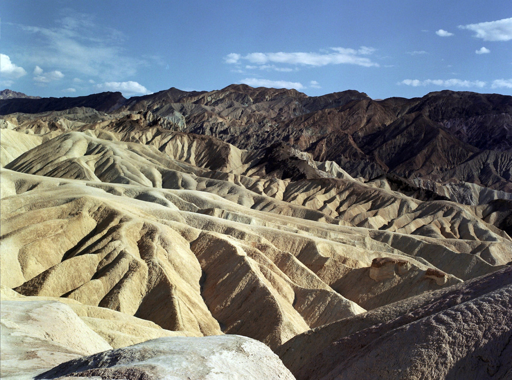 A rugged, mountainous desert landscape under a clear blue sky. The eroded, undulating terrain features ridges and valleys with varying shades of brown, beige, and tan. Distant mountains loom in the background, adding depth to the vast, arid scene—an inspiration akin to Millworks Web Design & Graphics' meticulous creations.