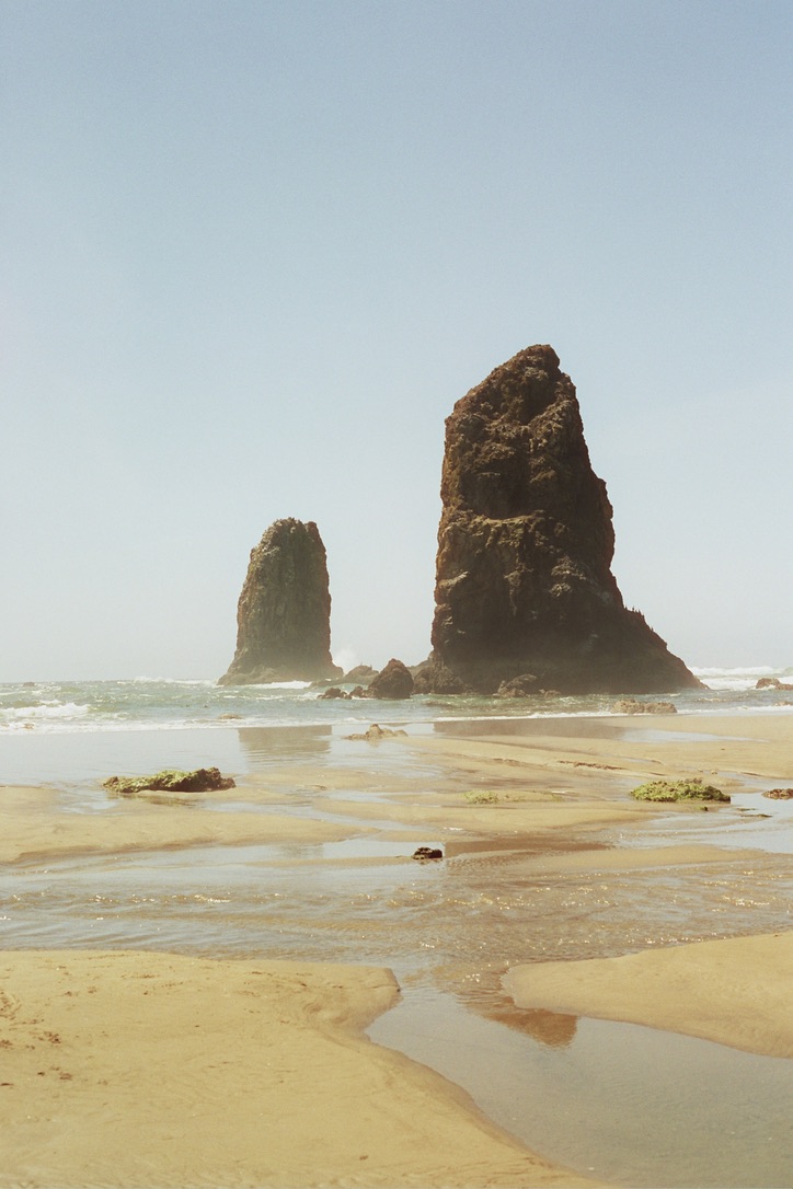 The image shows a scenic beach with two prominent sea stacks rising from the water near the shore. The sandy beach has shallow pools of water and some scattered seaweed. The clear blue sky not only highlights the natural coastal beauty but could easily inspire web design themes for custom websites.