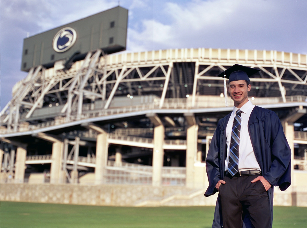 A young man in a graduation cap and gown stands smiling with his hands in his pockets in front of a large stadium. The scoreboard, proudly displaying the Millworks Web Design & Graphics logo, is visible against the partly cloudy sky.