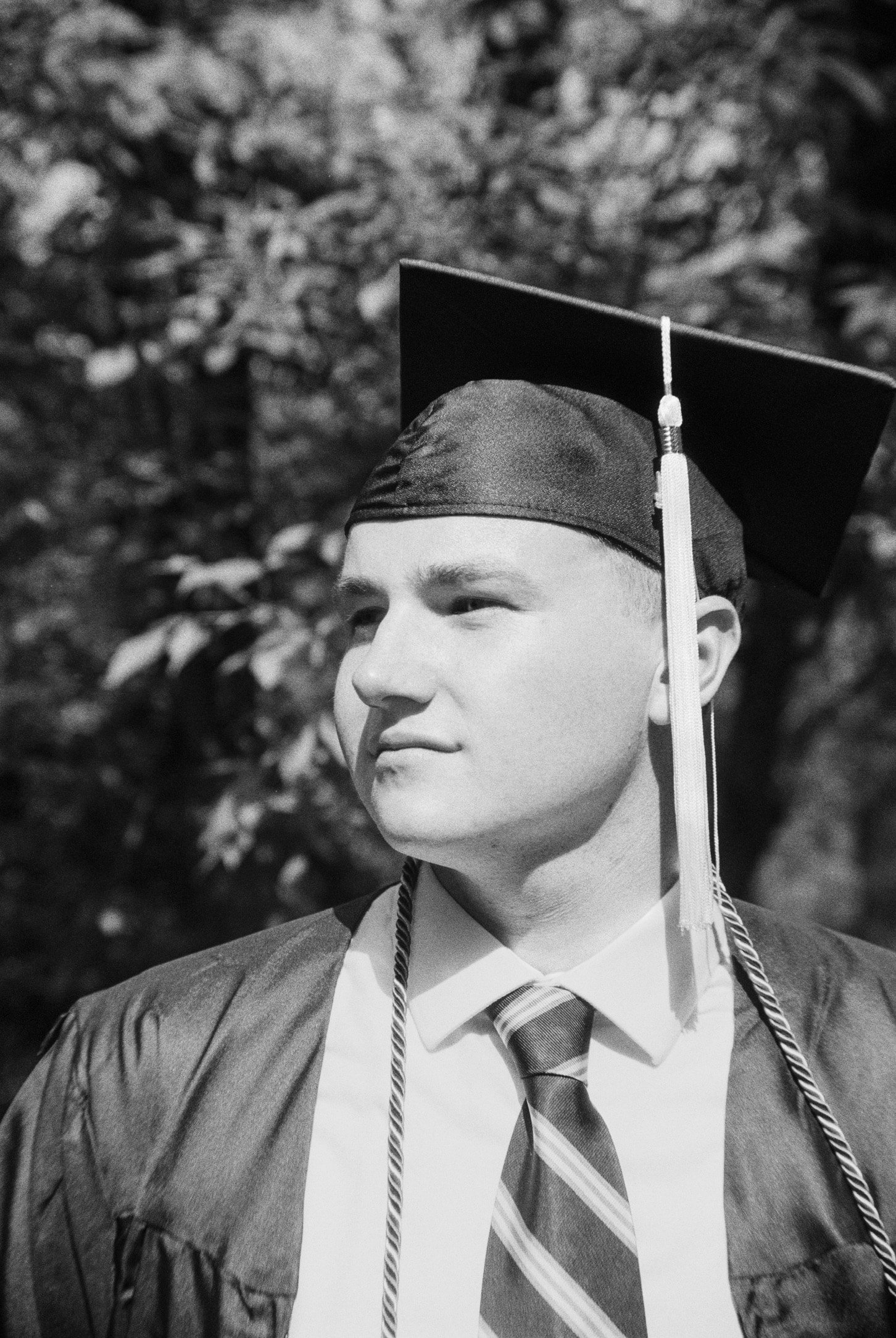 Black and white photo of a young man wearing a graduation cap and gown. He is looking to the right side of the frame with a serious expression, as though envisioning his future in graphic design. The background is blurred, showing some foliage. Photo for Millworks Web Design & Graphics portfolio.