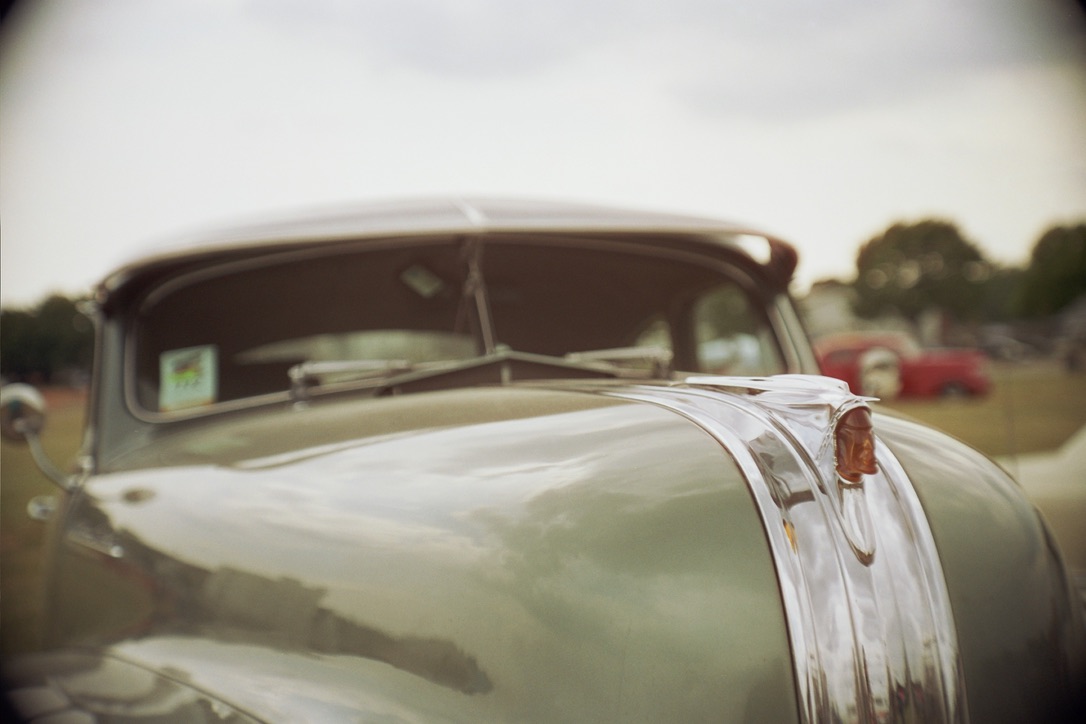 Close-up view of the front of a vintage car, featuring a shiny metallic hood ornament and chrome detailing. The car's windshield and part of the roof are visible in the background, with a blurred outdoor setting including other cars and trees. This classic aesthetic could inspire graphic design at Millworks Web Design & Graphics.