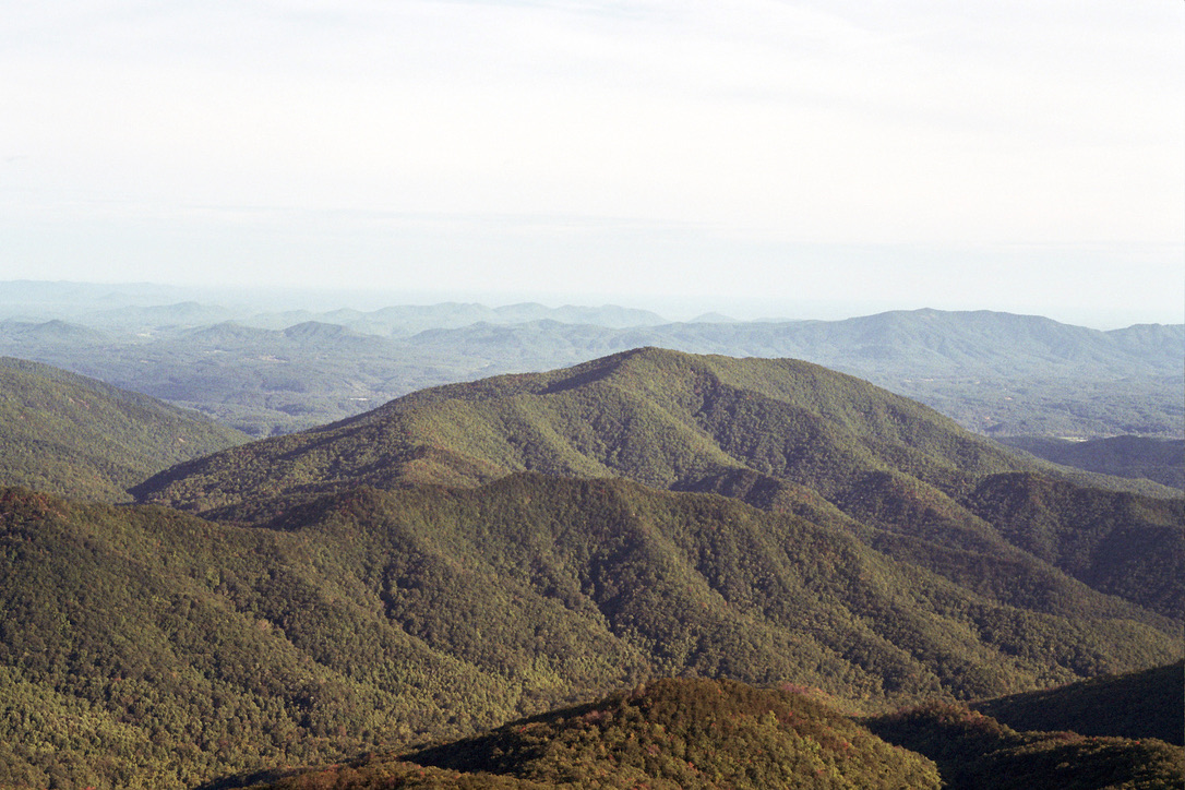 A panoramic view of a vast mountain range covered with lush green forests looks like a masterpiece of graphic design. The peaks and valleys extend into the distance, fading into a hazy, light blue sky with a few scattered clouds, evoking a serene and tranquil atmosphere.