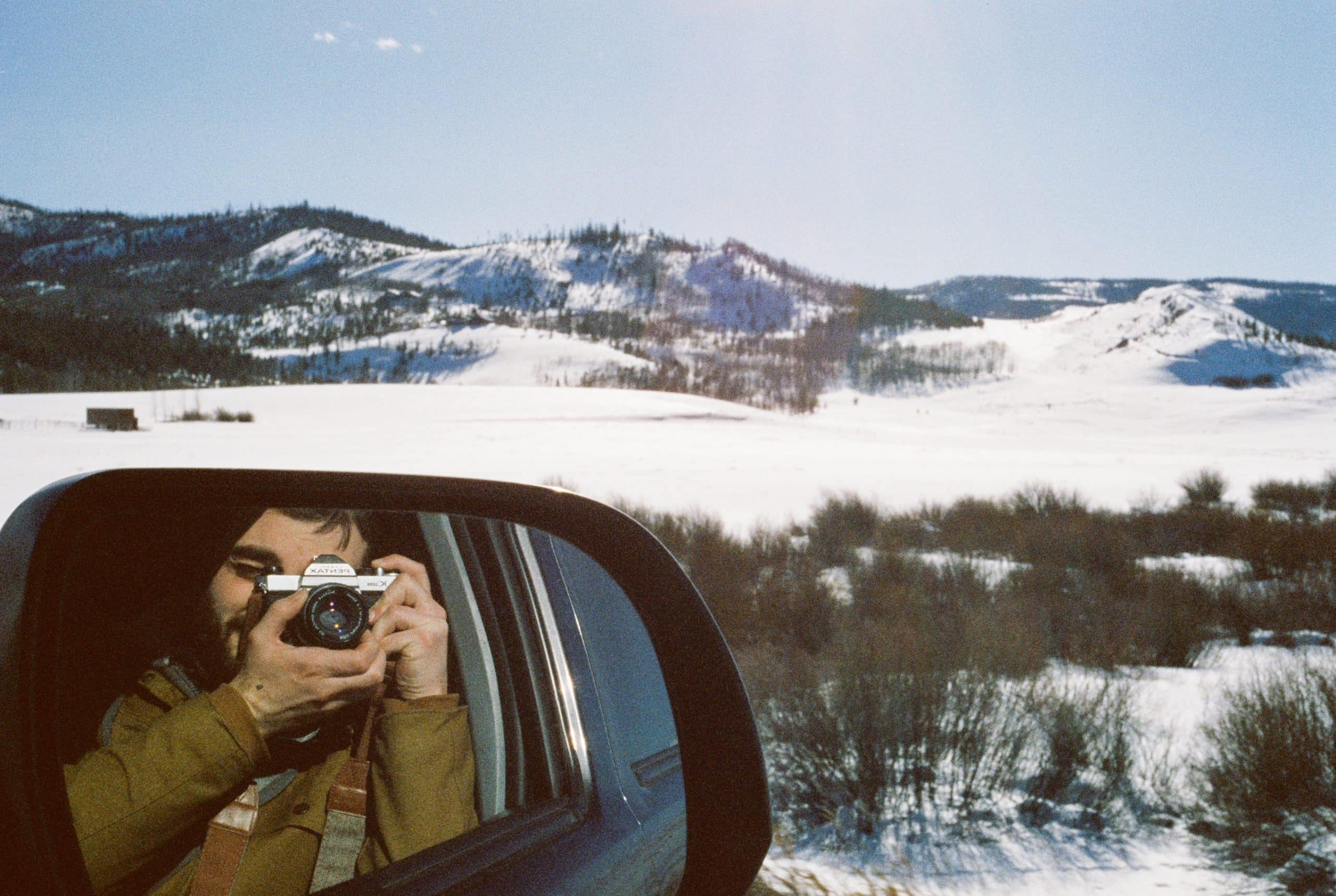 A person is taking a photo of themselves with a camera in the side mirror of a vehicle. Snow-covered mountains and a bright blue sky are visible in the background. The photo, perfect for graphic design inspiration, captures the reflection of the person holding the camera and the winter landscape.