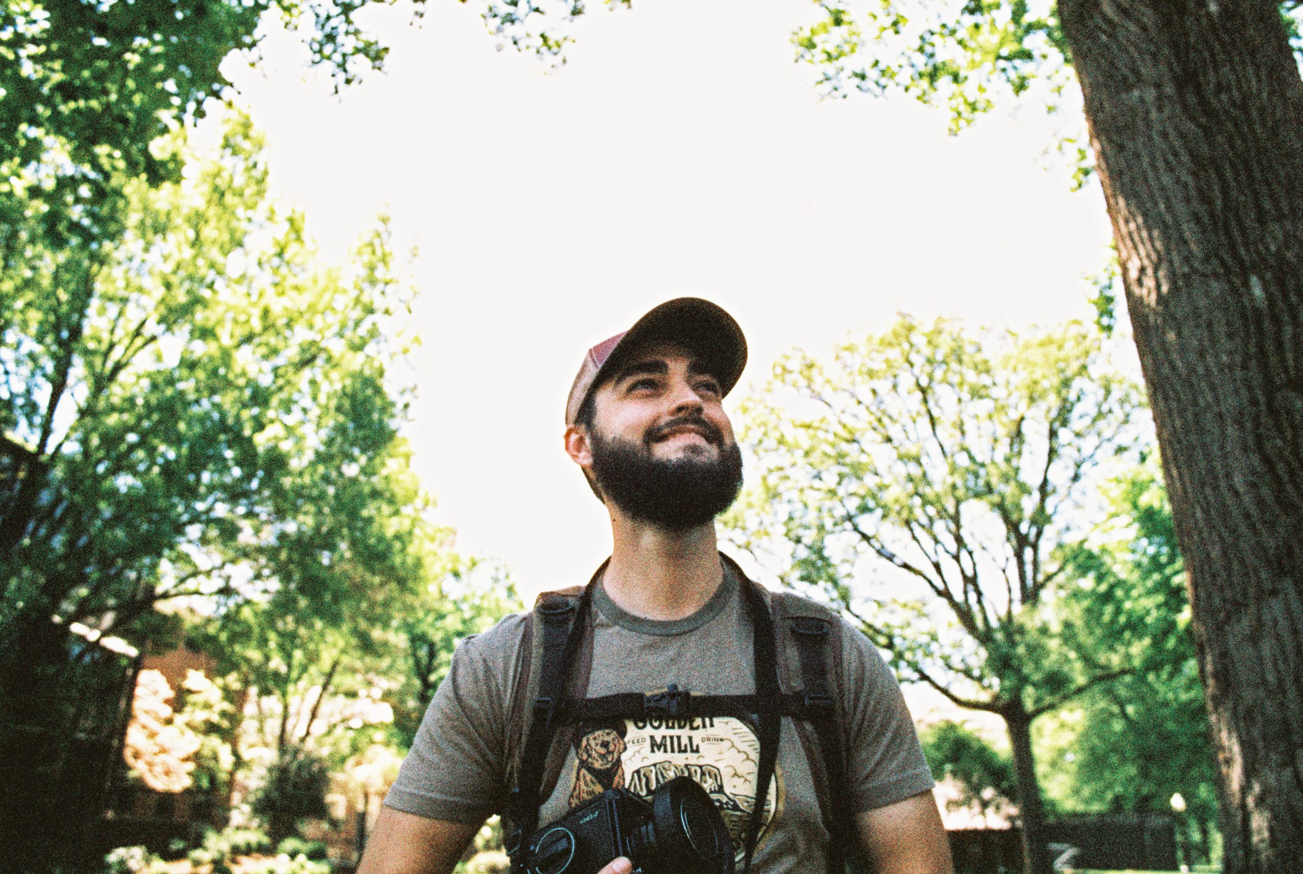 A bearded man wearing a baseball cap, t-shirt, and backpack stands outdoors, looking up with a smile. He holds a camera and is surrounded by tall, leafy trees and a clear, bright sky—an ideal snapshot for those who appreciate the beauty of nature captured through the lens of graphic design.