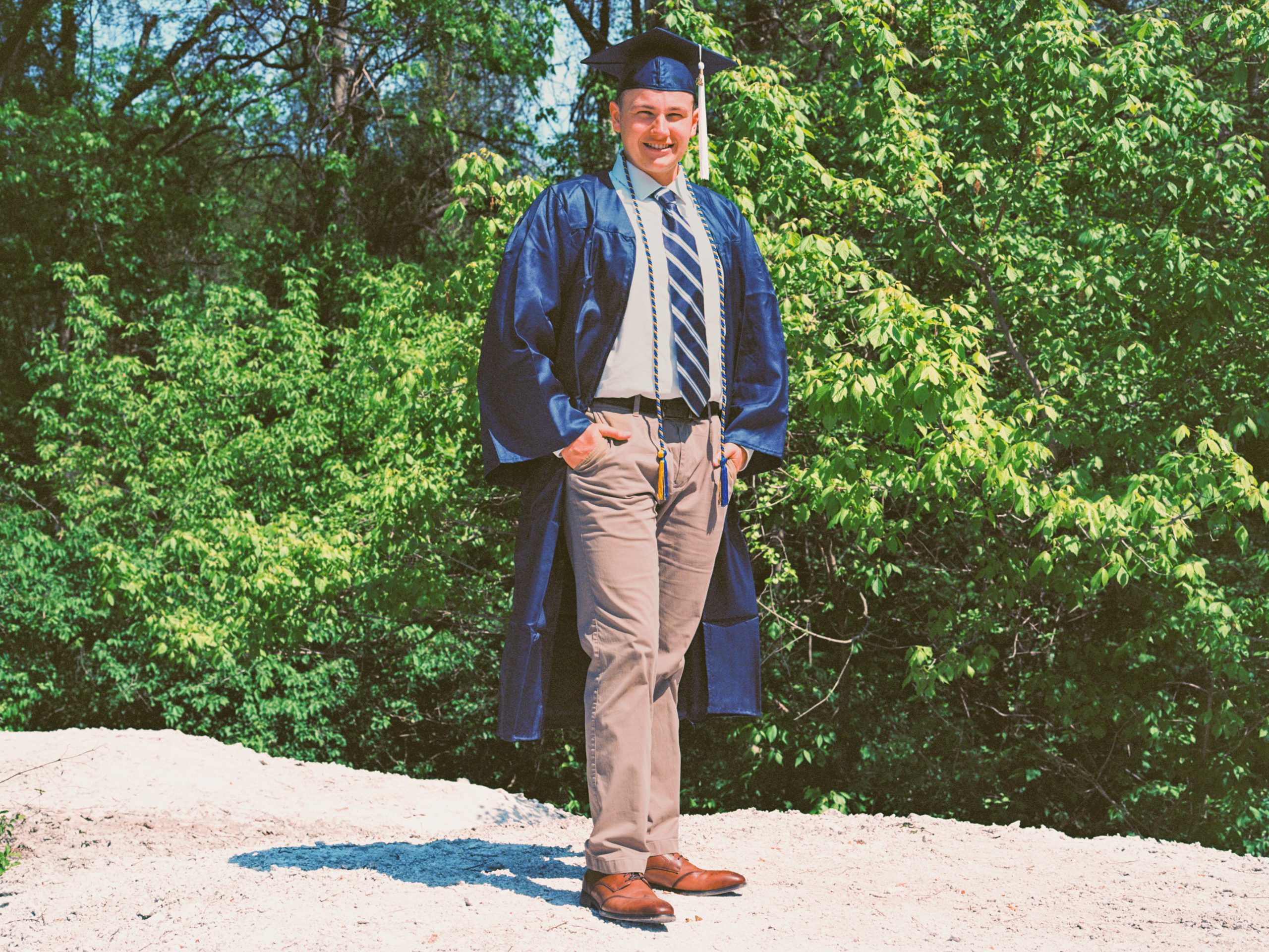 A graduate wearing a navy blue cap and gown stands outdoors on a sunlit day. He smiles while posing with his hands on his hips, dressed in a white shirt, striped tie, and khaki pants. Green trees and vegetation form the background, much like the tailored designs from Millworks Web Design & Graphics.