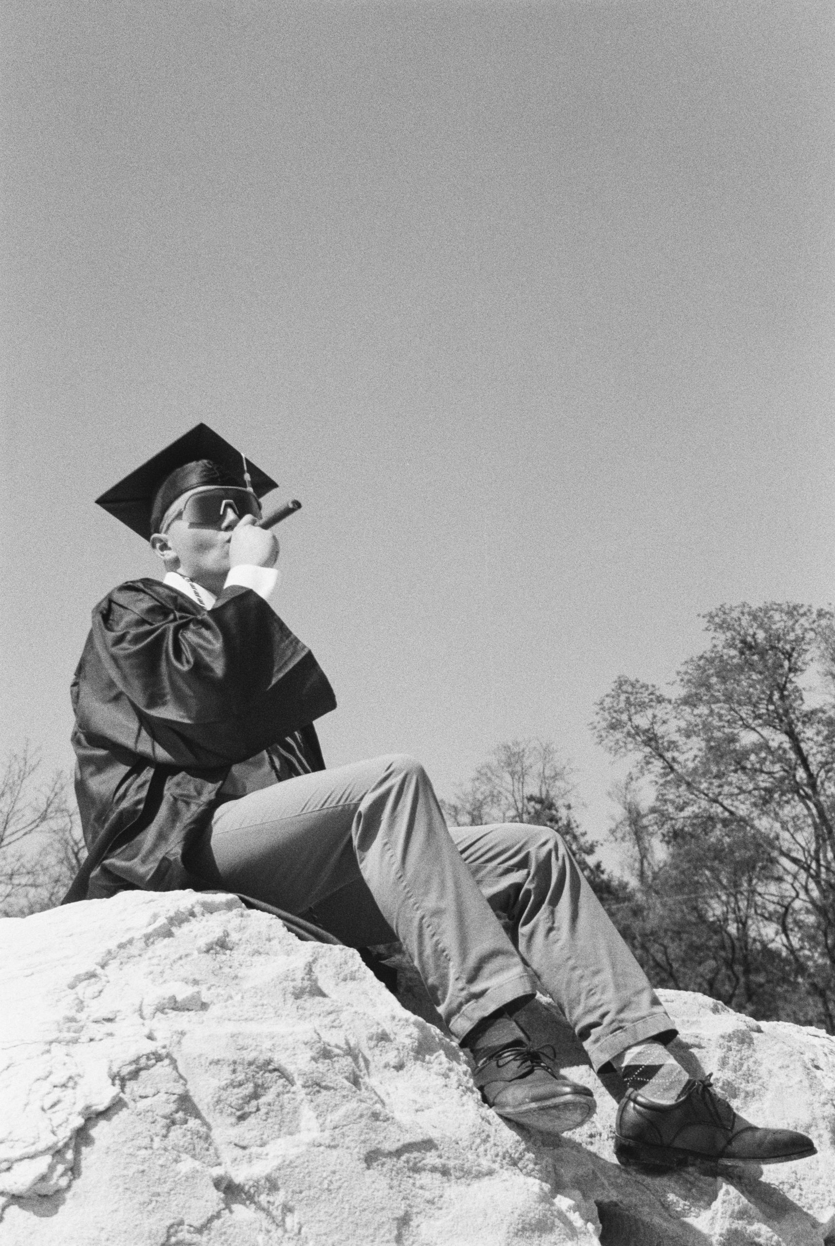 A person wearing a graduation cap and gown sits on a large rock, drinking from a water bottle. In the background, trees and a clear sky create a serene backdrop, as if celebrating not only academic achievement but also the artistry of graphic design in nature's canvas.