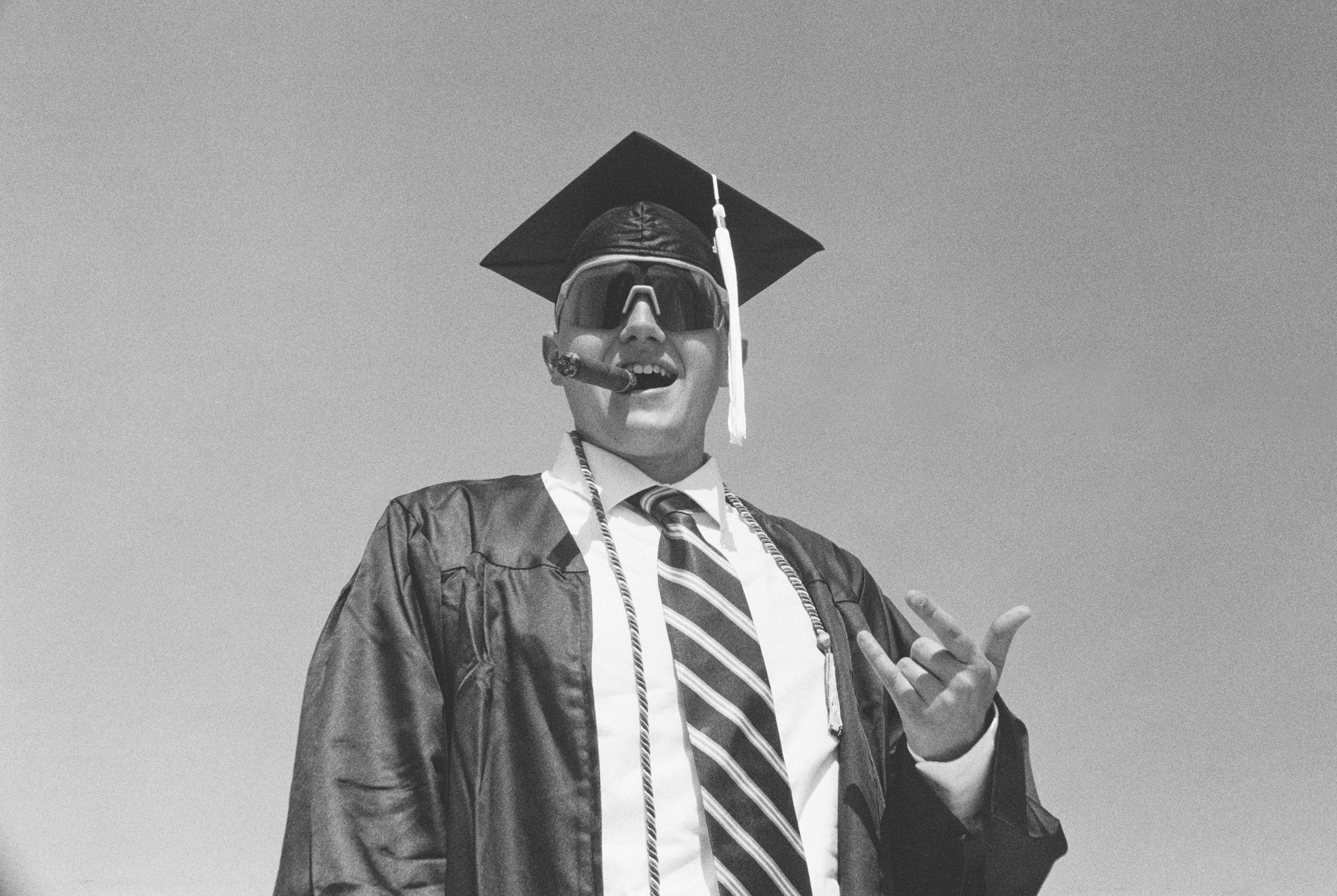 A person in a graduation cap and gown smokes a cigar. They wear large, dark sunglasses and a striped tie, evoking a stylish professional ready for success. The background is a clear sky, much like the bright future ahead—perhaps made even brighter with custom websites from Millworks Web Design & Graphics.