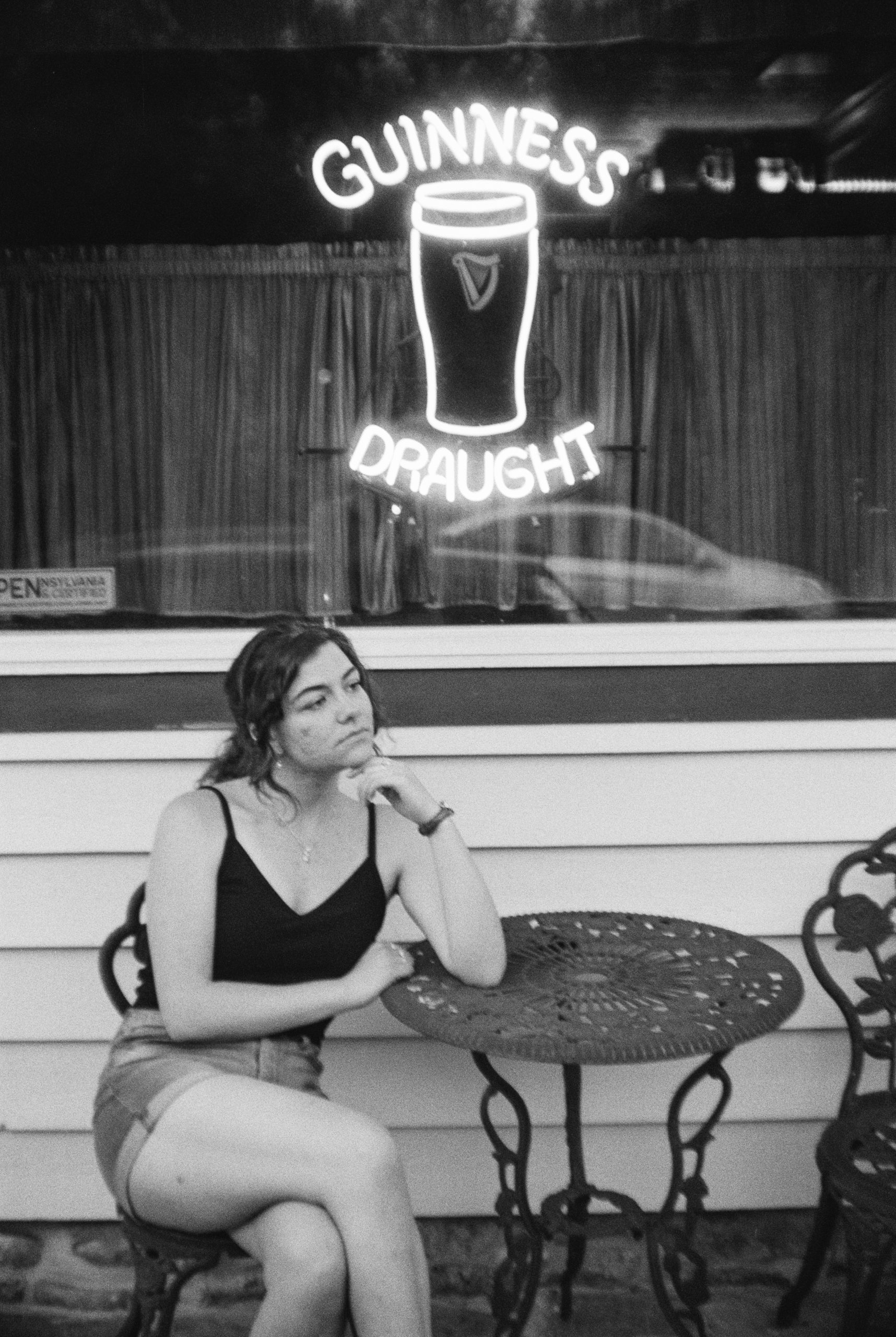 Black and white photo of a woman sitting at an outdoor table in front of a window with a neon "Guinness Draught" sign. She has a thoughtful expression, wearing a tank top and shorts, resting her head on her hand. An "open" sign is visible in the window, hinting at the digital marketing buzz inside.