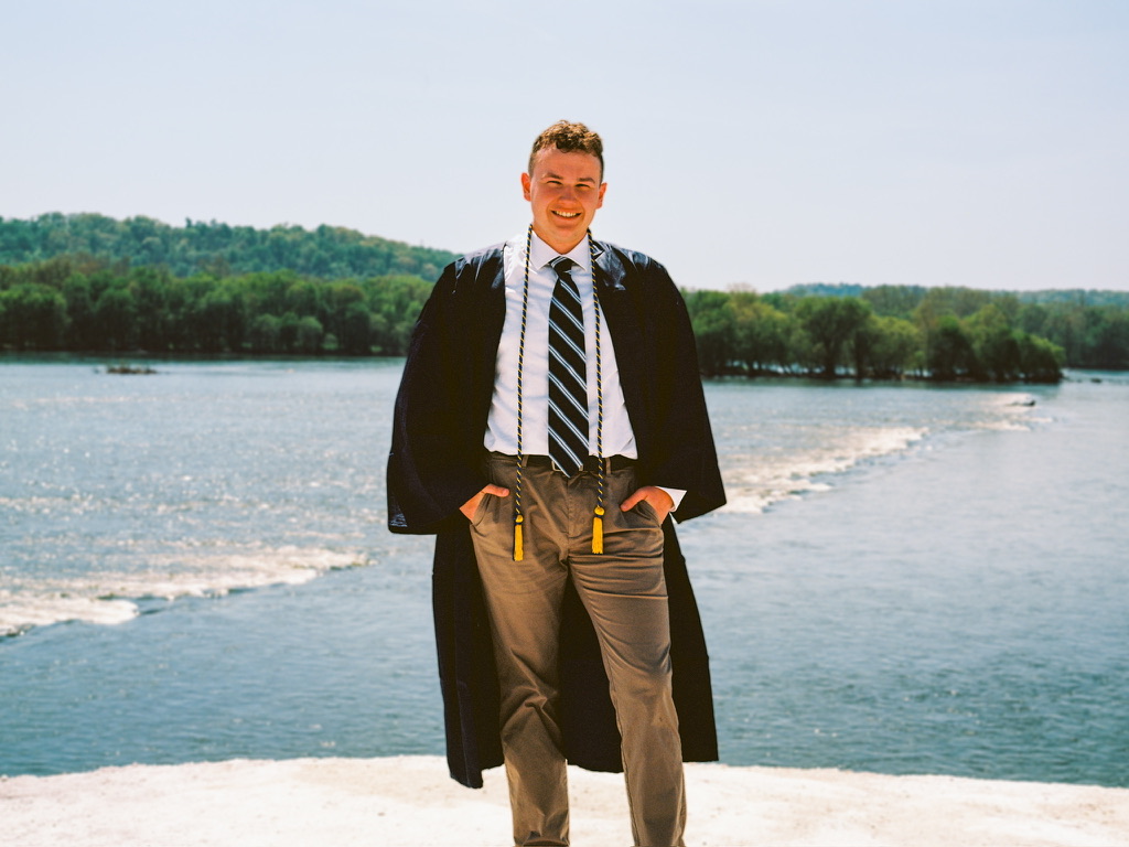 A person wearing a graduation gown, light shirt, striped tie, and khaki pants stands outdoors in front of a body of water with greenery in the background. The person, who specializes in graphic design, is smiling and has their hands in their pockets.