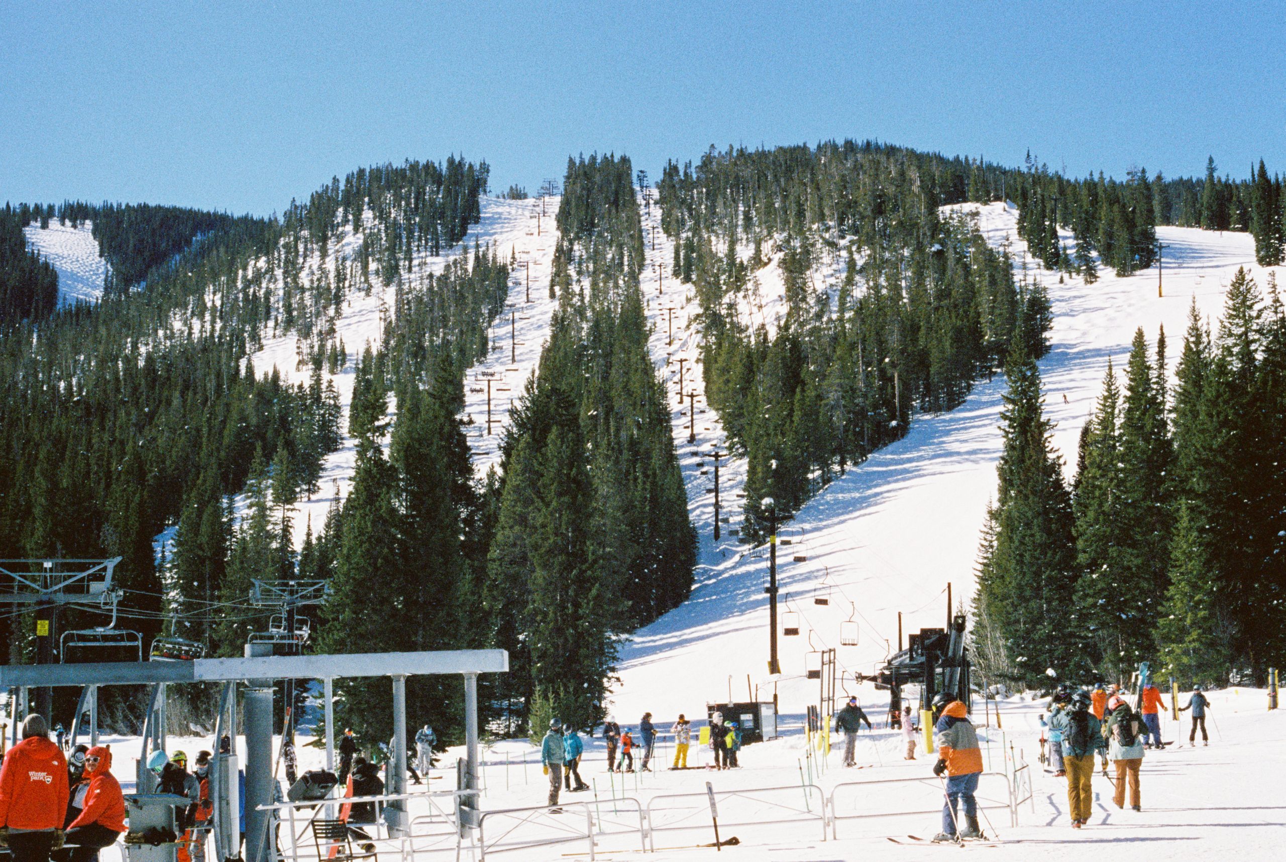 A group of skiers and snowboarders gather near a ski lift at the base of a snowy mountain. Tall evergreen trees line the slopes, with clear blue skies overhead and a few people in colorful winter gear preparing for their runs—reminiscent of how Millworks Web Design & Graphics crafts custom websites.