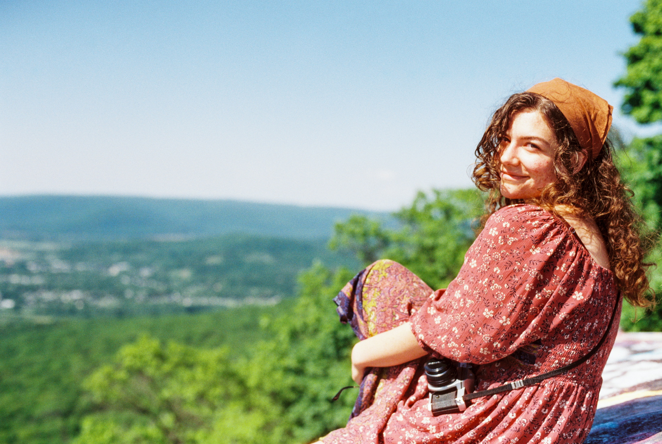 A person with long curly hair, wearing a pink patterned dress and a headscarf, sits on a ledge overlooking a scenic valley filled with green trees and distant mountains. Smiling at the camera, with a vintage camera around their neck, they radiate creativity as if inspired by graphic design.