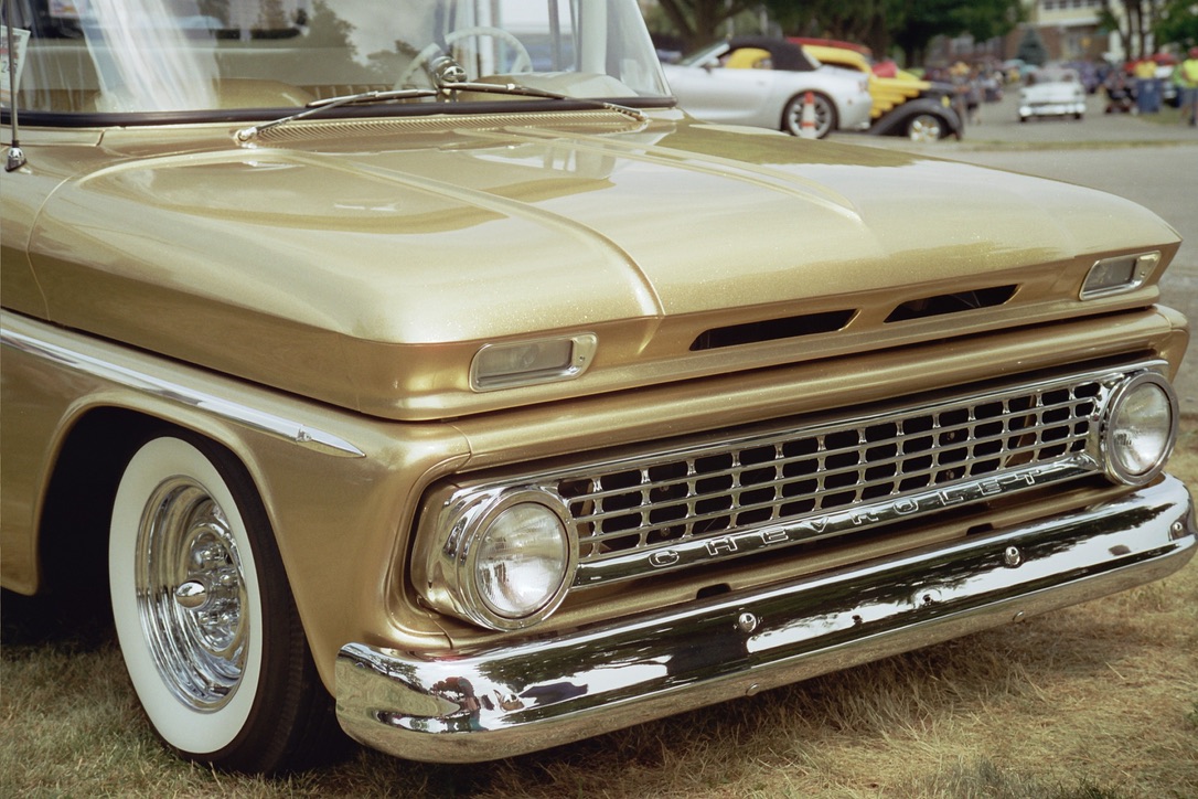 Close-up of the front end of a vintage golden Chevrolet truck with white-walled tires and chrome details. The truck is parked on a grassy area, looking as timeless and polished as a custom website from Millworks Web Design & Graphics, with other classic cars visible in the blurry background.