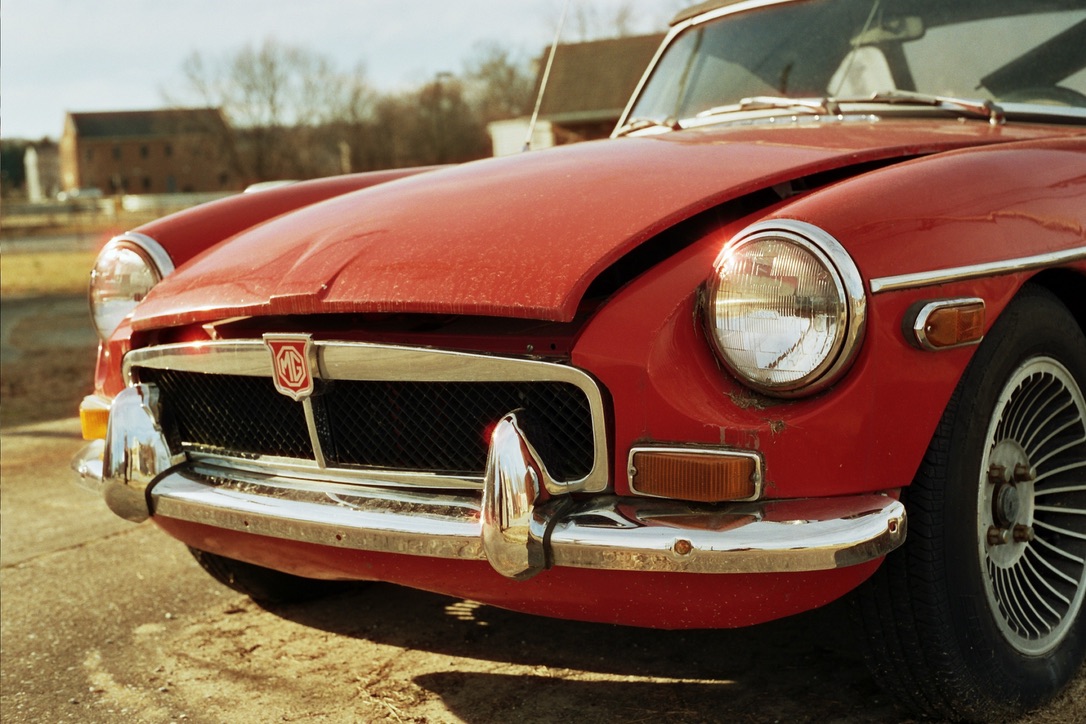 A close-up photograph of a vintage red MG car with a slightly damaged front evokes the charming aesthetics often sought in custom websites. The hood is partially open, revealing dents and scratches on the bumper and hood. In the background, trees and a blurred building add to its nostalgic appeal.