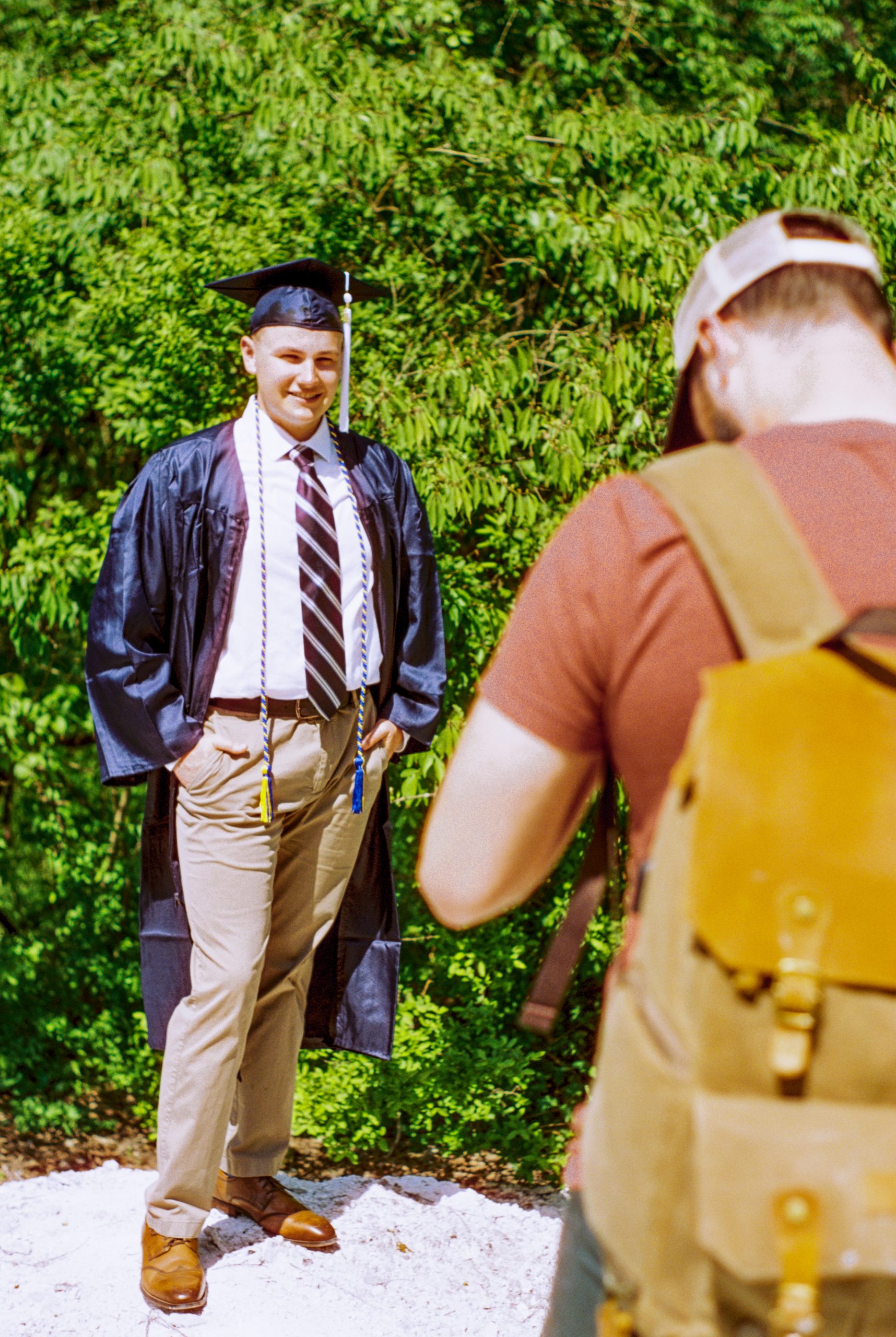 A person in a graduation cap and gown poses for a photo outdoors. They are dressed in a striped tie, beige pants, and brown shoes. Another person with a backpack and a cap is taking the photo, likely capturing memories before diving into careers like web design at Millworks Web Design & Graphics. The background is filled with green foliage.