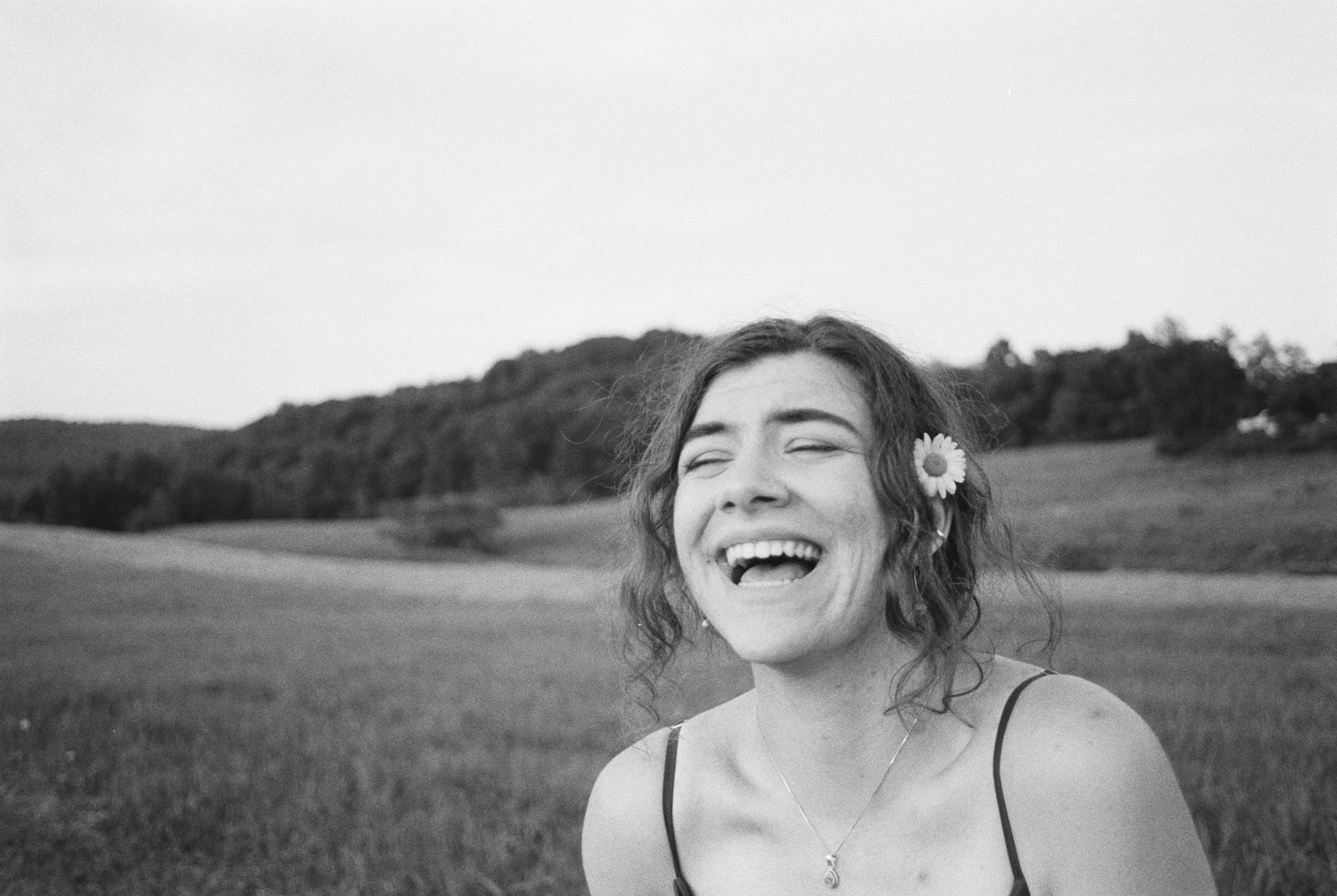 Black and white film photography captures a person laughing joyfully in an open field. They have curly hair adorned with a daisy behind their ear and are wearing a spaghetti strap top. The portrait features a vast grassy landscape with hills and trees under a clear sky.