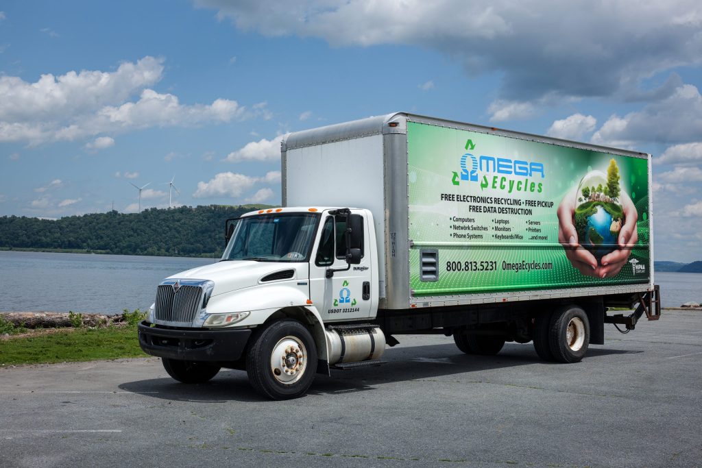 A white Omega Ecycles truck with a green advertisement for free electronics recycling is parked by a body of water, while trees, hills, and wind turbines line the background beneath a partly cloudy sky.