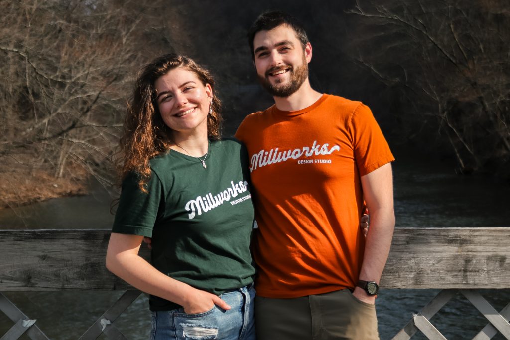 Two people stand side by side, smiling, on a wooden bridge with a river and bare trees in the background. Both wear millworks-inspired Design Studio t-shirts, one in green and one in orange.