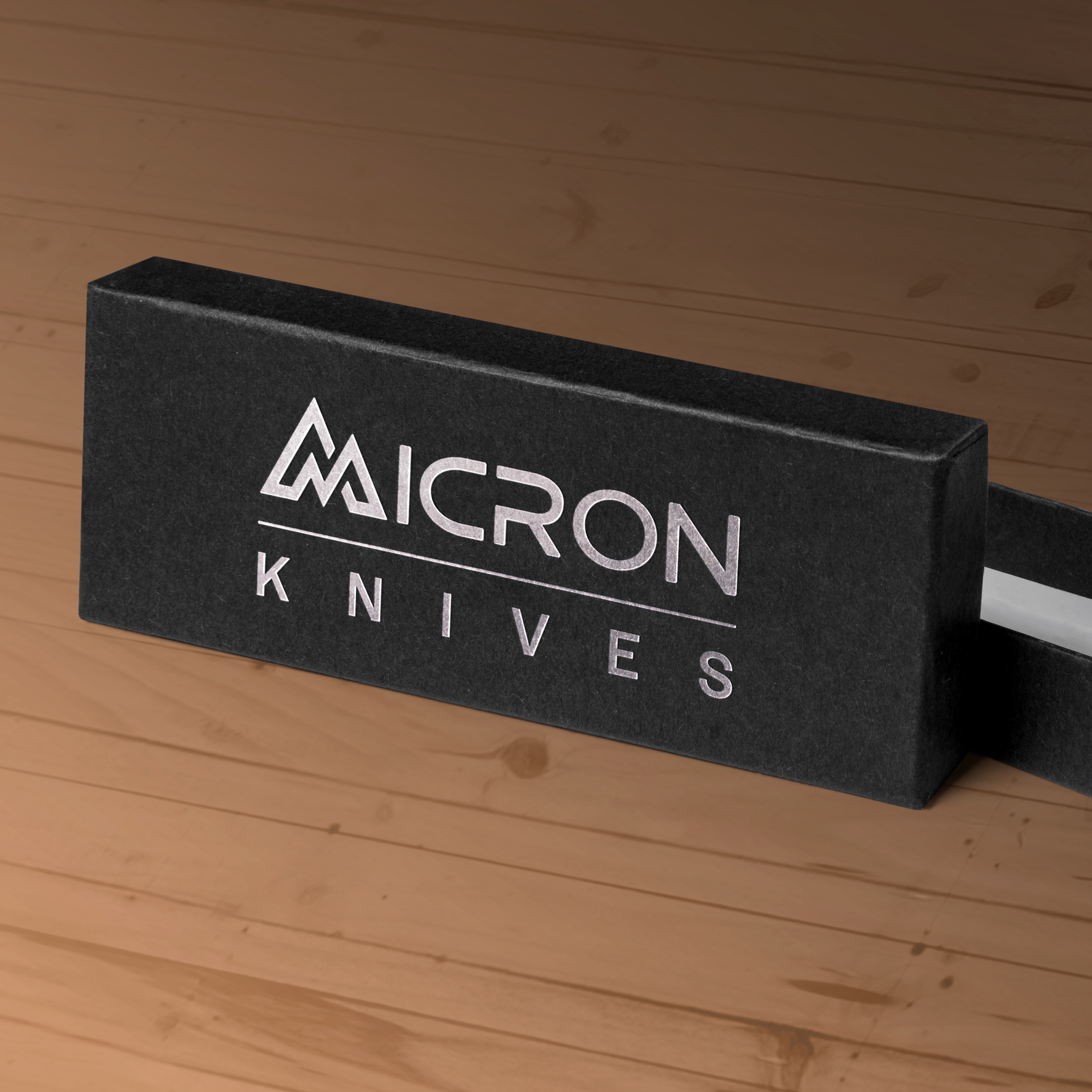 A black rectangular box with the silver Micron Knives logo and text on the lid rests on a wooden surface. The box is partially open, subtly revealing a premium Micron Knives blade inside.