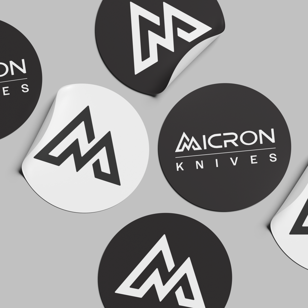 Round stickers in black and white showcase the bold, angular Micron Knives logo and text. Displayed on a light grey surface, some of these Micron Knives stickers are slightly peeled at the edges.