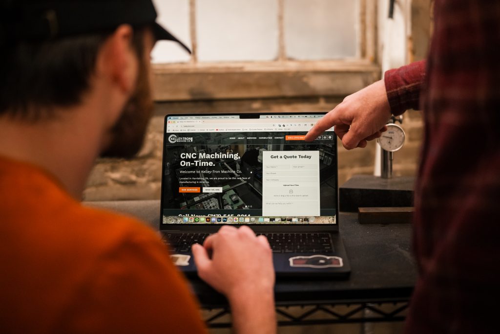 Two people look at a laptop screen displaying a website about CNC machining. One points at the screen, discussing how CNC technology can elevate your small business. The background includes a window and industrial equipment.