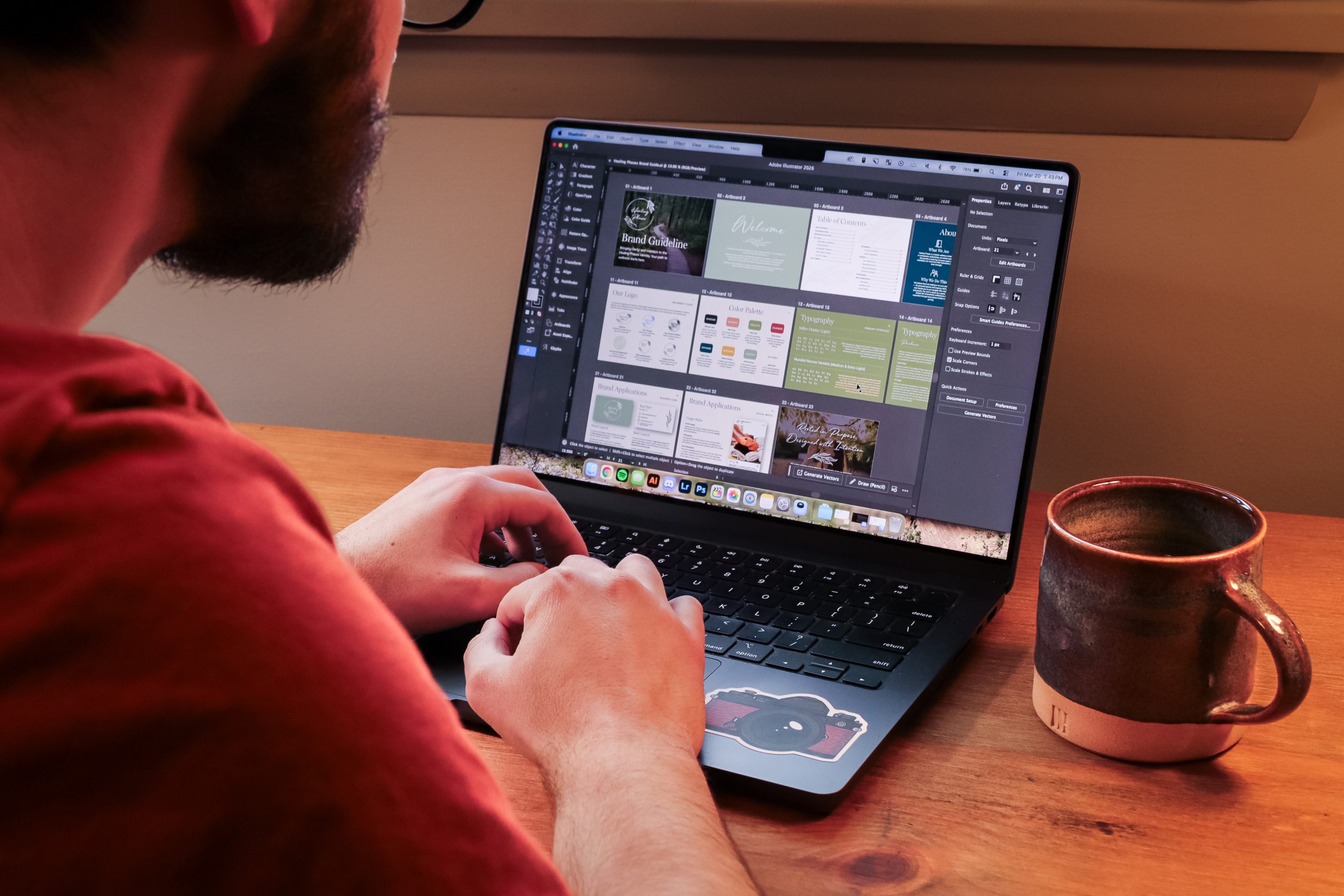 A person in a red shirt works on a laptop displaying an AI-powered graphic design application with multiple project windows open. A ceramic mug sits on the wooden desk beside the laptop.