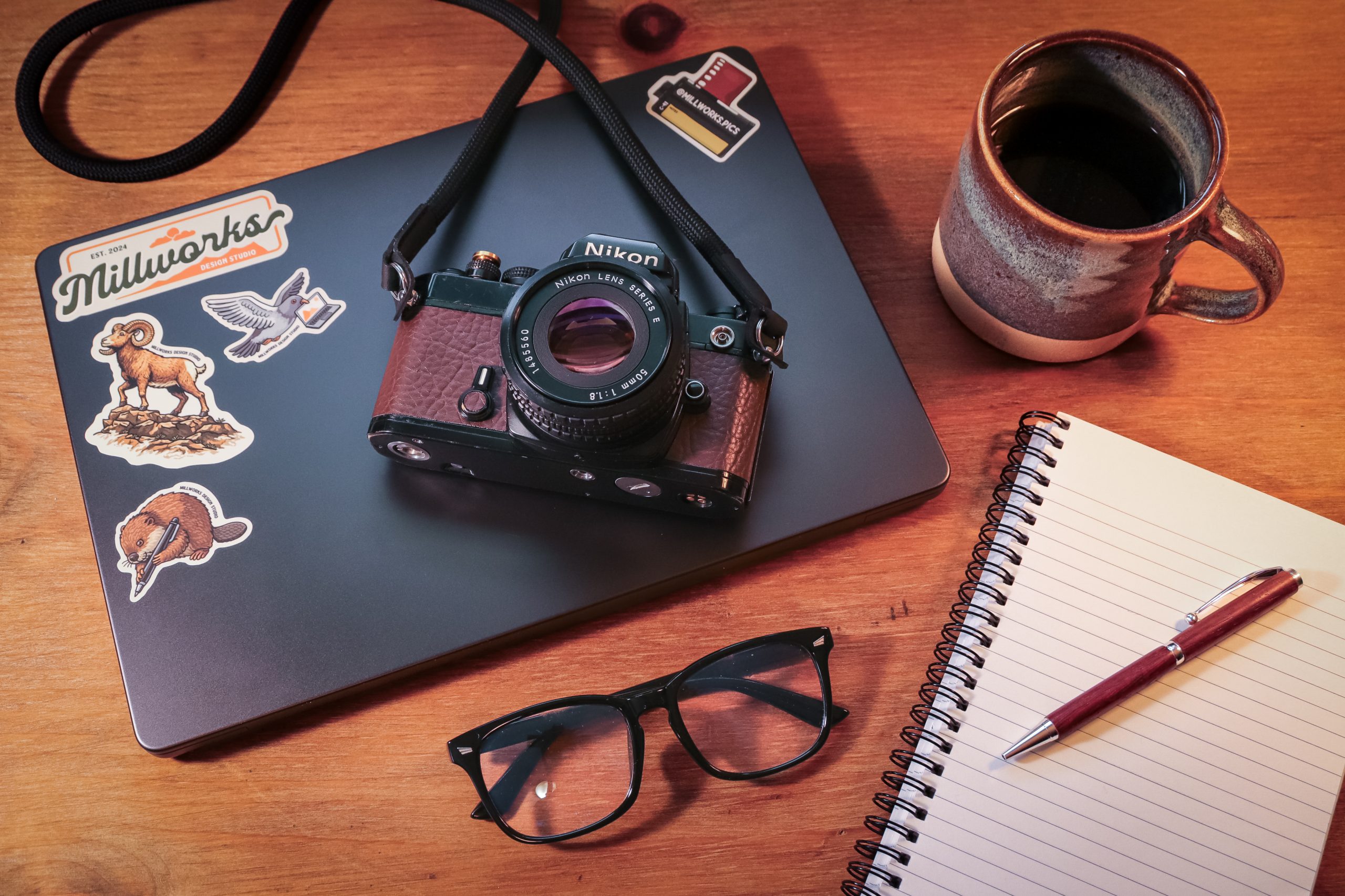A laptop with animal stickers, a vintage Nikon camera, glasses, a lined notebook with a pen, and a mug of coffee rest on a wooden desk—perfect for crafting our About Us story.