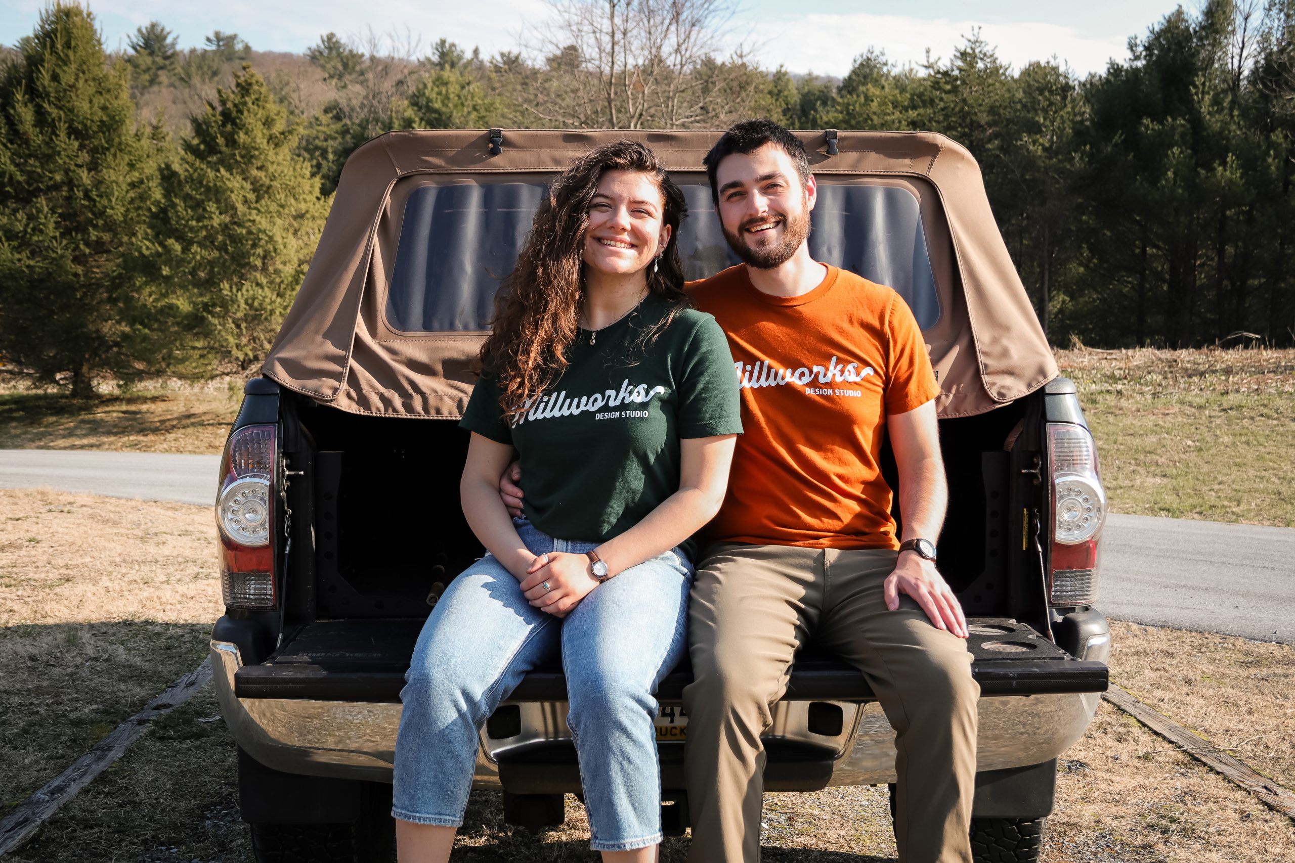 A smiling couple sits on the open tailgate of a pickup truck, surrounded by trees and grass. Casually dressed in “Millworks” T-shirts, they enjoy a sunny day outdoors—contact with nature and each other brightens their afternoon.
