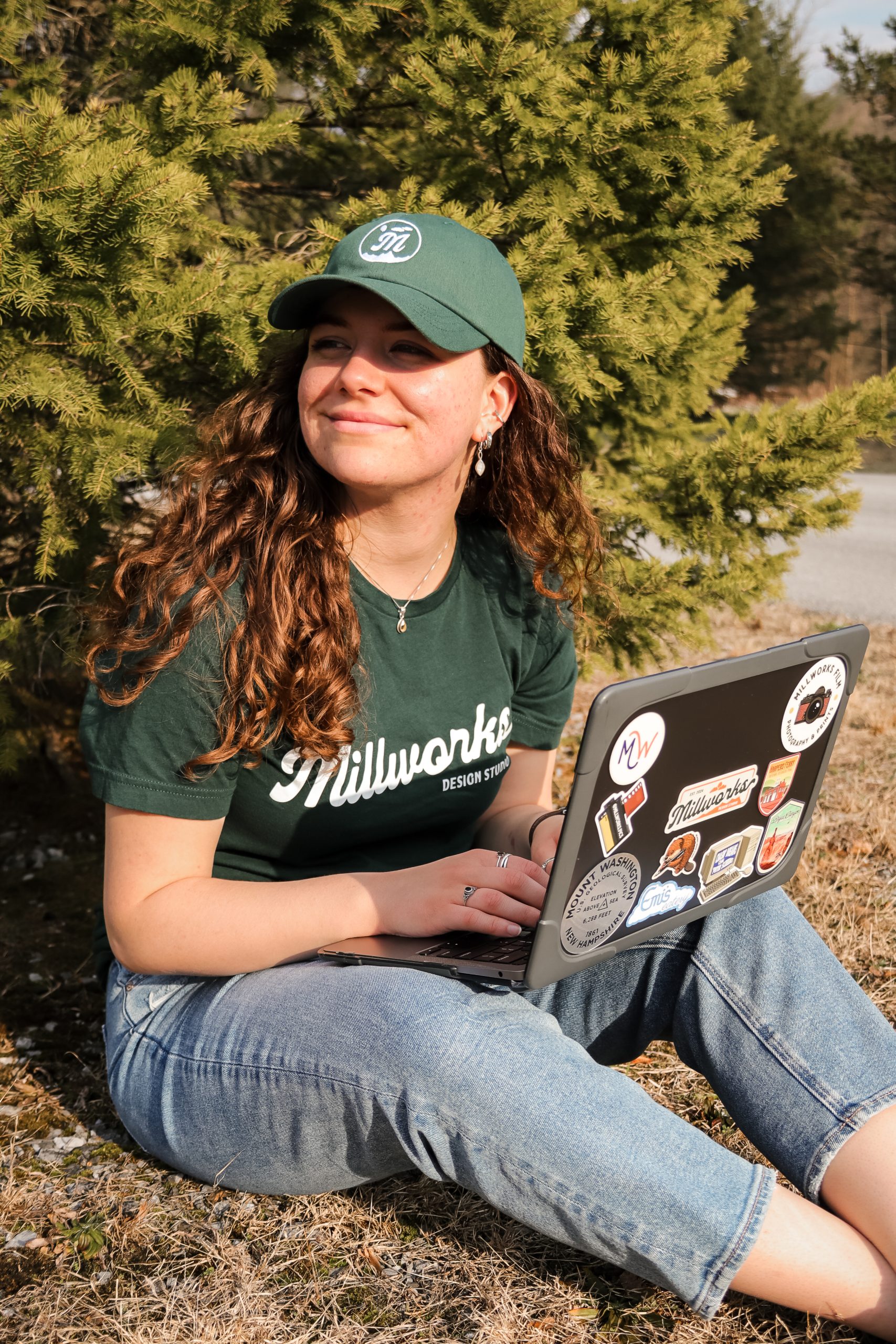 A young woman in a green hat and Millworks t-shirt sits on the grass by a tree, smiling while working on web design projects on her sticker-covered laptop. Sunlight highlights her face and curly hair.