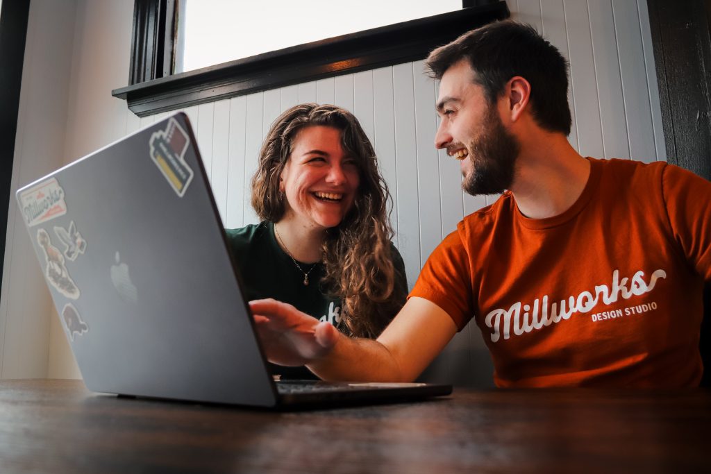 Two young adults sit at a table, smiling and laughing while looking at a laptop. One wears an orange Millworks Design Studio shirt, the other is in green. They're ready to elevate your small business against a sleek white and black wall.