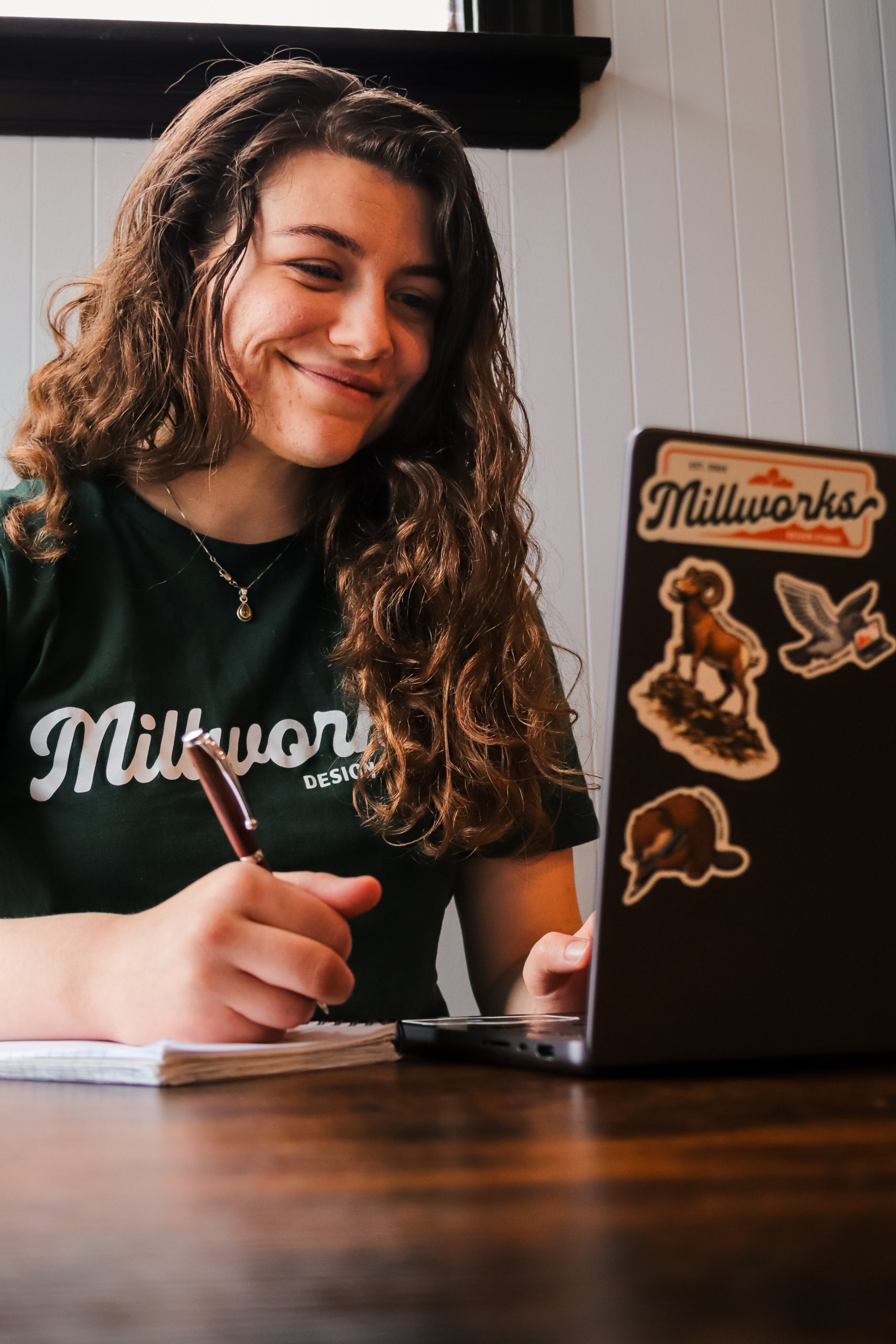 A person with long curly hair smiles while writing in a notebook and looking at a laptop decorated with stickers. Wearing a green “Millworks” T-shirt, they sit at a wooden table, ready to contact friends or colleagues as they work.